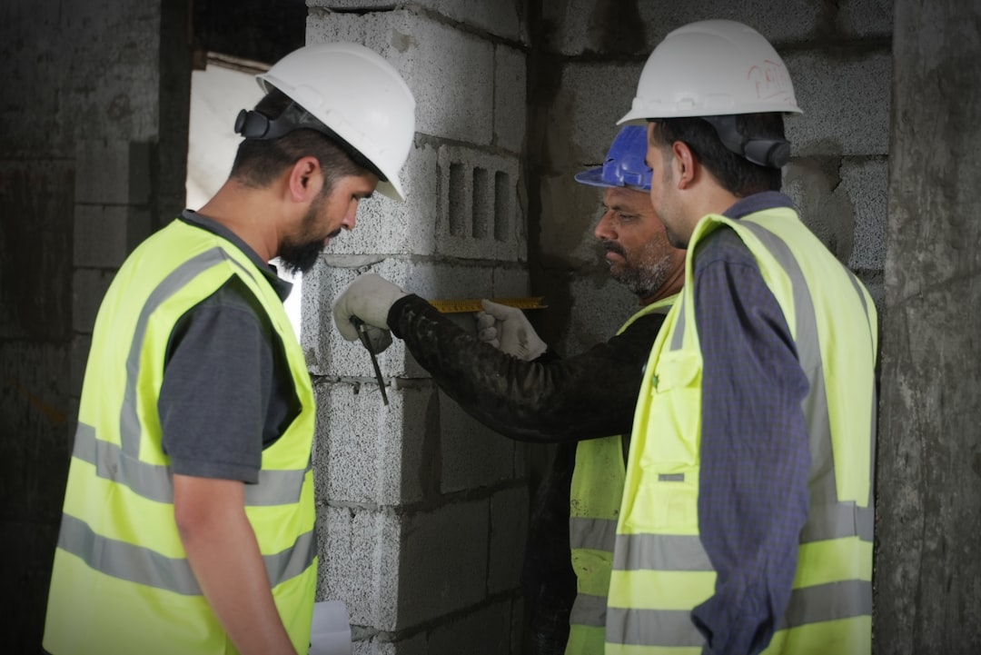 Three construction workers in high-visibility vests and hard hats measuring a concrete block wall